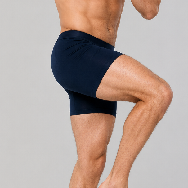 A shirtless man in dark navy athletic shorts is captured mid-jump against a plain light gray studio background.